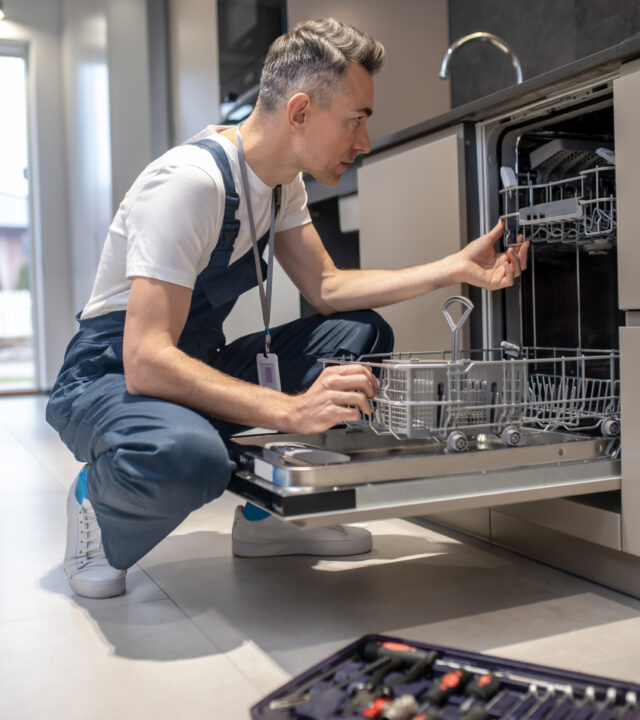 Profile of man crouching looking into open dishwasher Diagnostics of equipment. Profile of serious man in dark overalls with badge crouching attentively looking at open dishwasher in kitchen at home during day